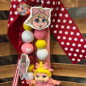 May include: A handmade lanyard featuring Miss Piggy. The lanyard has a red ribbon with white stars, a string of colorful beads, and a Miss Piggy figurine. The beads are white, pink, and yellow. The lanyard also has a small charm with a Miss Piggy image.