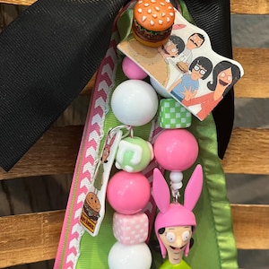 May include: A handmade keychain featuring a burger charm, cartoon character beads, and a small figurine of a character in a green dress and pink bunny ears. The keychain has a black ribbon and green and pink accents.