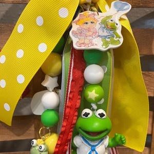 May include: A whimsical keychain featuring a yellow ribbon with white polka dots, a string of colorful beads, and a Kermit the Frog figurine. The keychain also includes a charm with two cartoon characters and a small frog figurine.