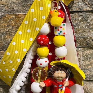 May include: A handmade keychain featuring a yellow polka dot ribbon, a yellow rubber duck, and a Cabbage Patch Kid figurine in a red coat holding a yellow umbrella. The keychain includes colorful beads and a gold-colored ring.