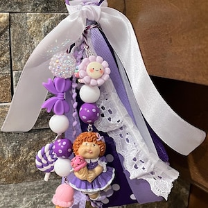 May include: A handmade charm featuring a Cabbage Patch Kid figurine holding an ice cream cone, with purple and white beads, ribbons, and a flower. The charm includes a white lace ribbon and a purple metal ring.
