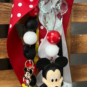 May include: A keychain featuring a large Mickey Mouse figure, a smaller Mickey Mouse charm, and various beads in red, white, and black. The accessory includes a red ribbon with white polka dots and a clear, faceted Mickey-shaped bead attached to a metal clip.