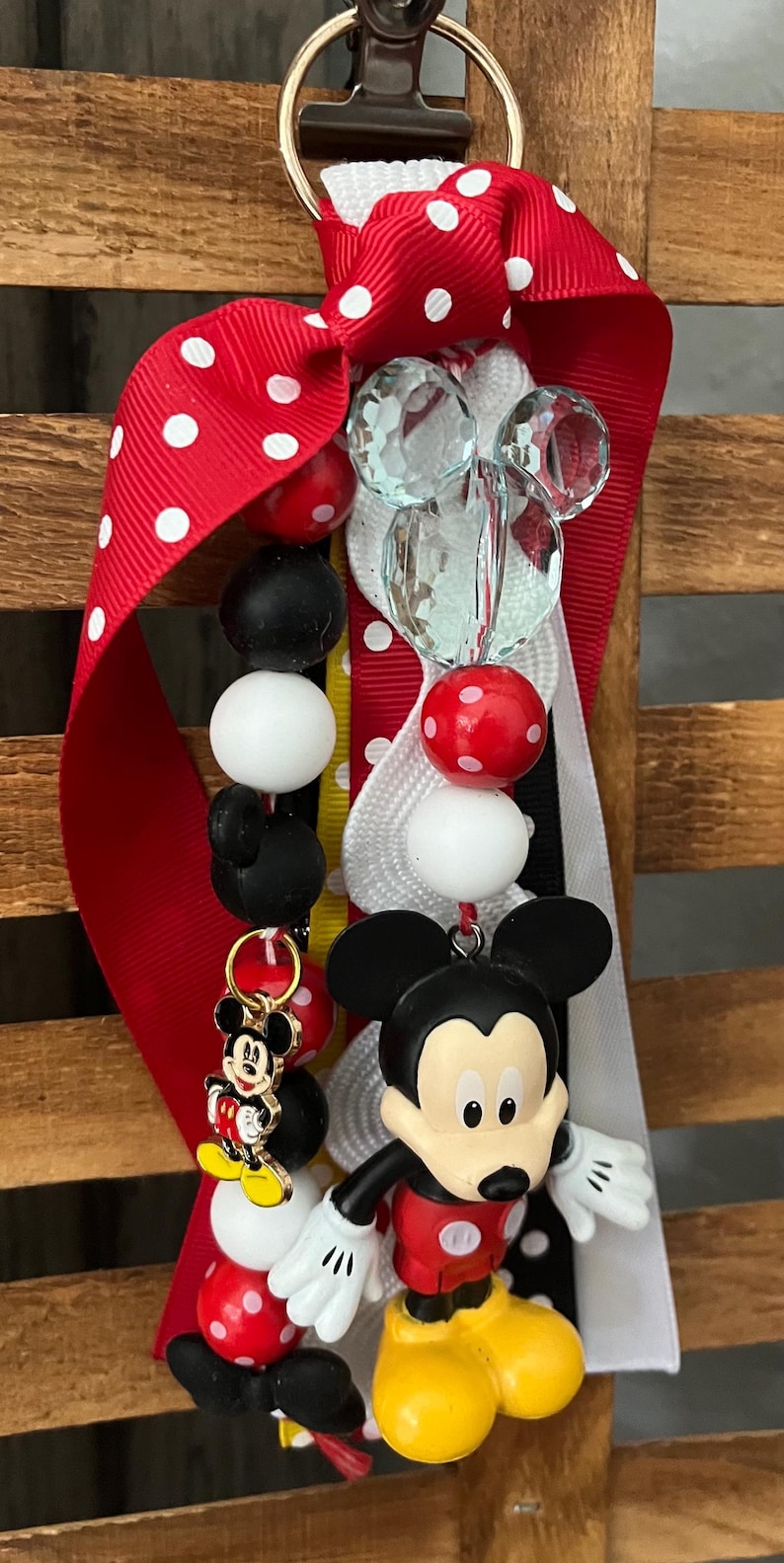 May include: A keychain featuring a large Mickey Mouse figure, a smaller Mickey Mouse charm, and various beads in red, white, and black. The accessory includes a red ribbon with white polka dots and a clear, faceted Mickey-shaped bead attached to a metal clip.