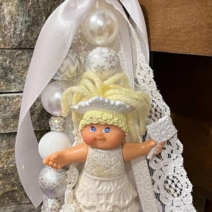 May include: A white and silver beaded keychain featuring a doll in a white dress with blonde hair. The keychain includes white ribbon, lace, and decorative beads in various sizes and textures. A gold-colored ring is at the top.