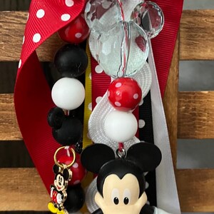 May include: A keychain featuring a Mickey Mouse figurine, a small Mickey Mouse charm, and various beads in red, white, and black. The beads include solid colors, polka-dot patterns, and a clear faceted bead. Red and white polka-dot ribbons are attached.
