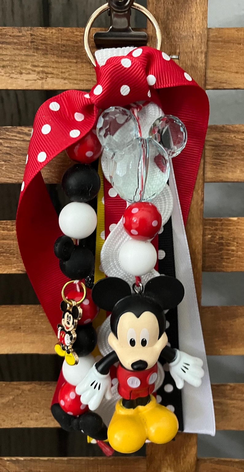 May include: A keychain featuring a Mickey Mouse figurine, a small Mickey Mouse charm, and various beads in red, white, and black. The beads include solid colors, polka-dot patterns, and a clear faceted bead. Red and white polka-dot ribbons are attached.