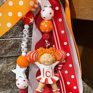 May include: A cheerleader-themed keychain with a Cabbage Patch Kids doll charm. The keychain features orange and white polka dot ribbon, red and white striped ribbon, and beads spelling out "CHEER". The doll is wearing a cheerleading uniform.