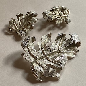 May include: A silver tone leaf brooch and matching earrings. The brooch is large and features a textured leaf design with a smaller leaf detail. The earrings are smaller and feature a similar leaf design.