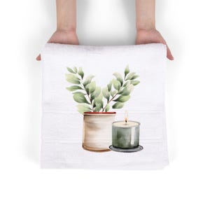 May include: White hand towel with a watercolor design of a potted plant and a lit candle. The plant has green leaves and is in a beige pot. The candle is in a clear glass holder with a gray base.