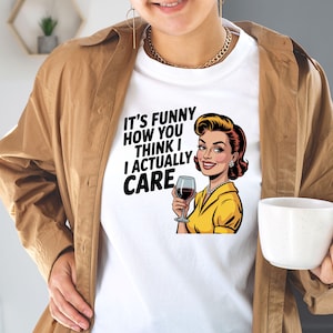 May include: White t-shirt with the text "IT'S FUNNY HOW YOU THINK I ACTUALLY CARE" and a retro illustration of a woman holding a wine glass. The woman is wearing a yellow top and has red hair.