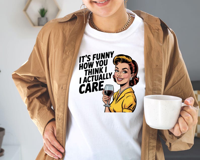 May include: White t-shirt with the text "IT'S FUNNY HOW YOU THINK I ACTUALLY CARE" and a retro illustration of a woman holding a wine glass. The woman is wearing a yellow top and has red hair.