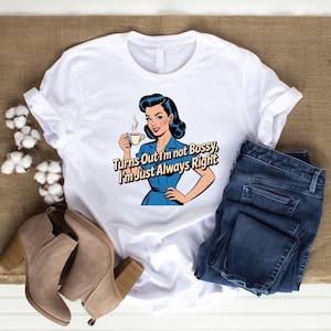 May include: White t-shirt with a retro illustration of a woman holding a coffee cup. The text on the shirt reads "Turns Out I'm not Bossy, I'm Just Always Right." The shirt is paired with blue jeans and tan ankle boots.