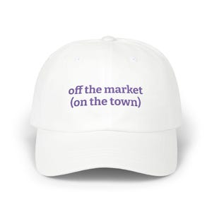 May include: A white baseball cap with a curved peak. The cap displays the text "off the market (on the town)" in purple lettering. This hat is a classic design, suitable for casual wear.