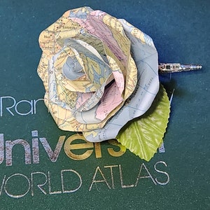 May include: A paper rose made from a world atlas map, with a green leaf and a silver pin. The rose is on a green book with the words "Rand McNally World Atlas" printed on the cover.