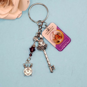 May include: A silver key charm with a white rabbit, a purple crystal bead, and a silver clock charm. The charm is attached to a silver key ring with a purple and white book charm that says "Alice's Adventures in Wonderland" by Lewis Carroll.