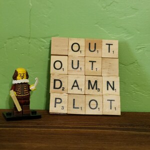 May include: A yellow Lego figure of William Shakespeare stands in front of a set of wooden letter tiles spelling out "OUT DAMN PLOT".