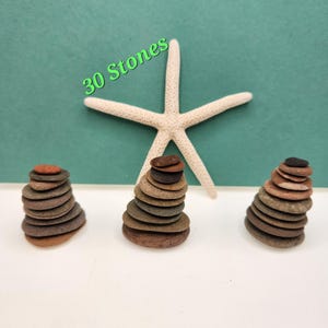 May include: Three stacks of small, flat stones in shades of brown and gray, with a white starfish and the text "30 Stones" on a teal background. The stones are arranged in a balanced, stacked formation.