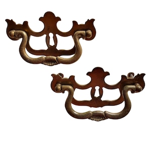 May include: Two antique brass drawer pulls with a decorative design. Each pull features a curved handle and an ornate backplate with a keyhole detail. The pulls are made of a warm, aged brass.