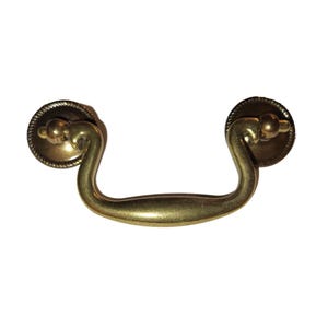 May include: A brass-toned drawer pull with a curved handle and circular backplates. The backplates have a rope-like border and a small, rounded knob at the center. The handle curves gracefully between the backplates.
