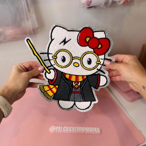 May include: A white Hello Kitty patch with a red bow, round glasses, and a lightning bolt scar. The patch is dressed in a black robe and a Gryffindor scarf, holding a wand. The patch has a silver glitter border and the text "@MI.CUSTOMPRINTS".