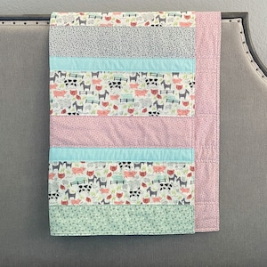May include: A multi-colored baby quilt with a farm animal print. The quilt has stripes of pink, gray, light blue, and green fabric. The farm animal print features pigs, cows, and horses.
