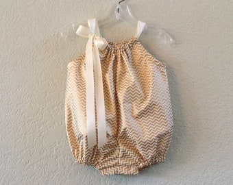 May include: A baby onesie with a chevron pattern in gold and white. The onesie has a white satin bow on the shoulder.