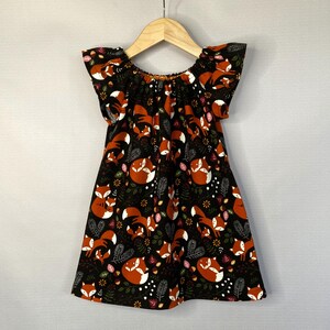 May include: A black dress with a repeating pattern of orange foxes, white accents, and green foliage. The dress has short sleeves and is hanging on a wooden hanger. The dress is designed for children.