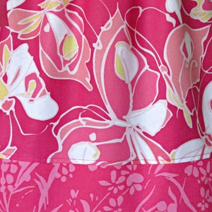 May include: Close-up of a vibrant pink fabric with a floral pattern. The design features large, stylized flowers in shades of pink, white, and yellow. The lower section has a border with a smaller floral print. The fabric appears to be a dress or skirt.