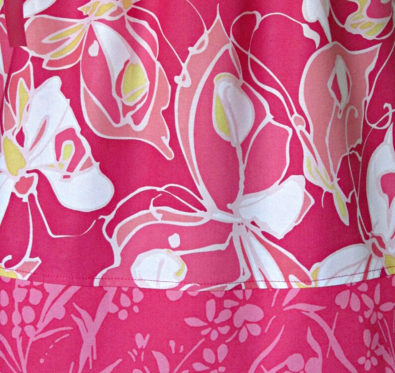 May include: Close-up of a vibrant pink fabric with a floral pattern. The design features large, stylized flowers in shades of pink, white, and yellow. The lower section has a border with a smaller floral print. The fabric appears to be a dress or skirt.