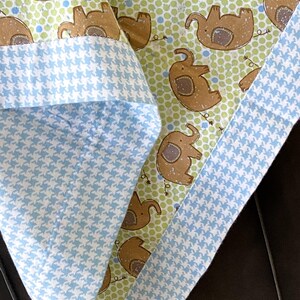 May include: A baby blanket with a blue and white houndstooth pattern on one side and a green background with brown elephants on the other side.