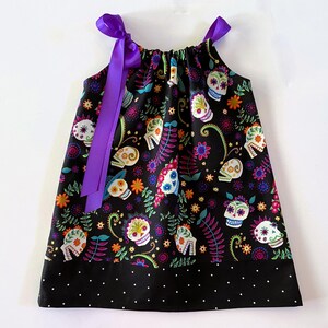 May include: A black sleeveless dress with a colorful Day of the Dead sugar skull print and purple ribbon straps. The dress has a black polka dot trim.