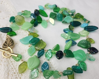 May include: A green glass leaf necklace with a gold clasp. The necklace is made of various shades of green glass leaves.