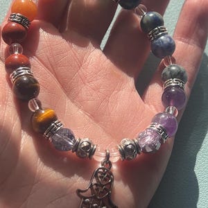 May include: A rainbow chakra bracelet with a silver hamsa charm. The bracelet features beads in red, orange, yellow, green, blue, indigo, and violet.