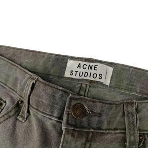 Puede incluir: Primer plano de unos vaqueros con estampado de camuflaje. Los vaqueros tienen cierre de bot&oacute;n y trabillas para el cintur&oacute;n. Se ve una etiqueta blanca con el texto "ACNE STUDIOS".