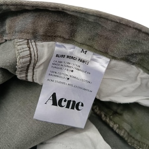 Puede incluir: Primer plano de una etiqueta de prenda con la marca "Acne" en texto negro. La etiqueta tambi&eacute;n incluye "BLISS MERCI PAW13", la talla "M" y detalles sobre el origen y el material de la prenda. La prenda tiene un estampado de camuflaje.