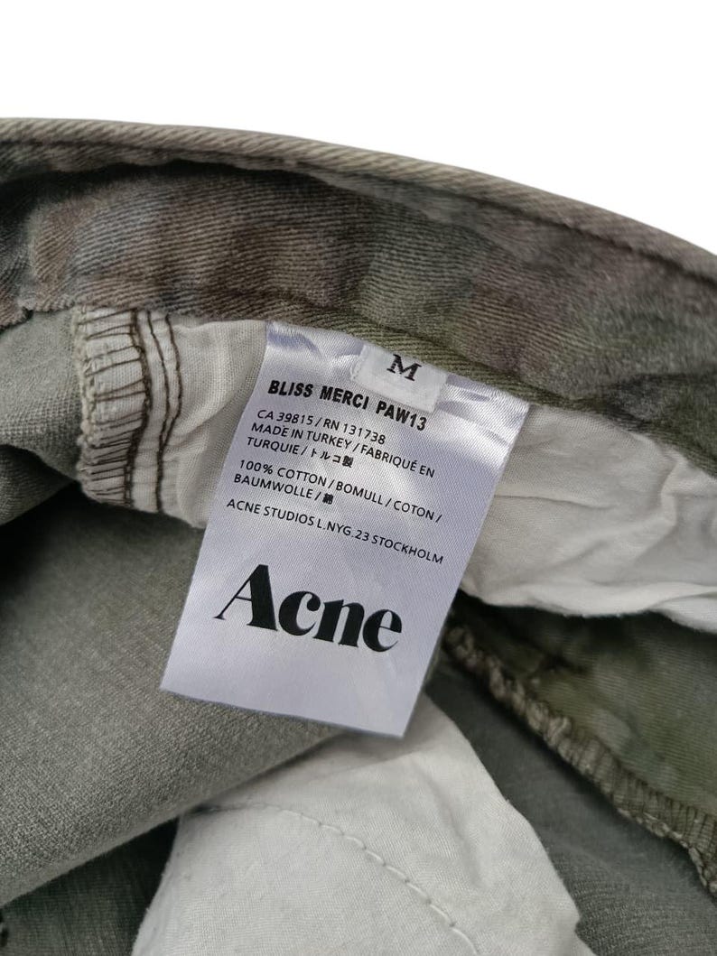 Puede incluir: Primer plano de una etiqueta de prenda con la marca "Acne" en texto negro. La etiqueta tambi&eacute;n incluye "BLISS MERCI PAW13", la talla "M" y detalles sobre el origen y el material de la prenda. La prenda tiene un estampado de camuflaje.