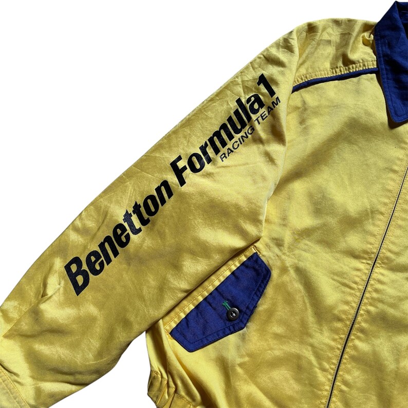 May include: Yellow jacket with a blue pocket and a black "Benetton Formula 1 Racing Team" logo on the sleeve.