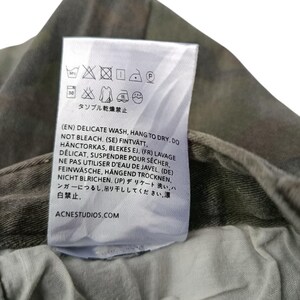 Puede incluir: Primer plano de una etiqueta de ropa blanca con instrucciones de lavado y el texto "ACNESTUDIOS.COM". La etiqueta est&aacute; adherida a una tela con estampado de camuflaje. La etiqueta incluye s&iacute;mbolos para lavado delicado, colgar para secar y no usar lej&iacute;a.