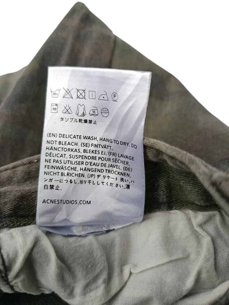 Puede incluir: Primer plano de una etiqueta de ropa blanca con instrucciones de lavado y el texto "ACNESTUDIOS.COM". La etiqueta est&aacute; adherida a una tela con estampado de camuflaje. La etiqueta incluye s&iacute;mbolos para lavado delicado, colgar para secar y no usar lej&iacute;a.