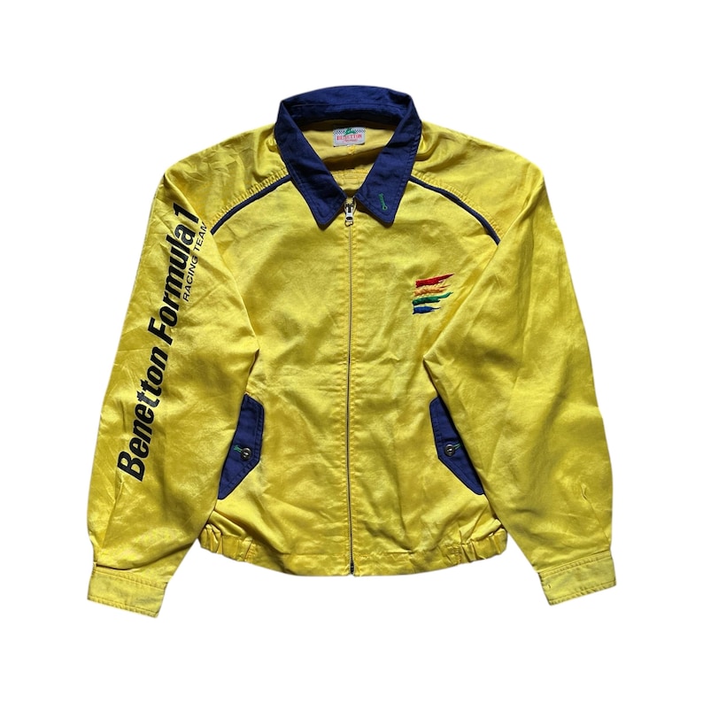 May include: A yellow jacket with blue trim and the text "Benetton Formula 1 Racing Team" on the left sleeve. The jacket has a zipper closure and a rainbow stripe logo on the right side.