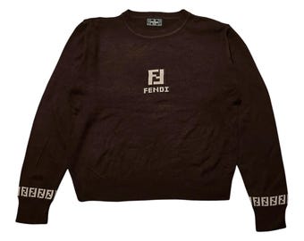 May include: A brown sweater with the Fendi logo in the center and on the cuffs. The sweater has a crew neck and long sleeves. The Fendi logo is in a white, blocky font. The sweater is a solid color with no other visible design elements.