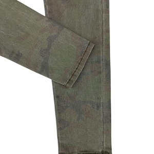 Puede incluir: Primer plano de unos vaqueros con estampado de camuflaje verde oliva. La tela presenta una mezcla de tonos verdes, marrones y negros. Los vaqueros est&aacute;n doblados, mostrando el dobladillo y la textura del material. El estilo general es informal y pr&aacute;ctico.