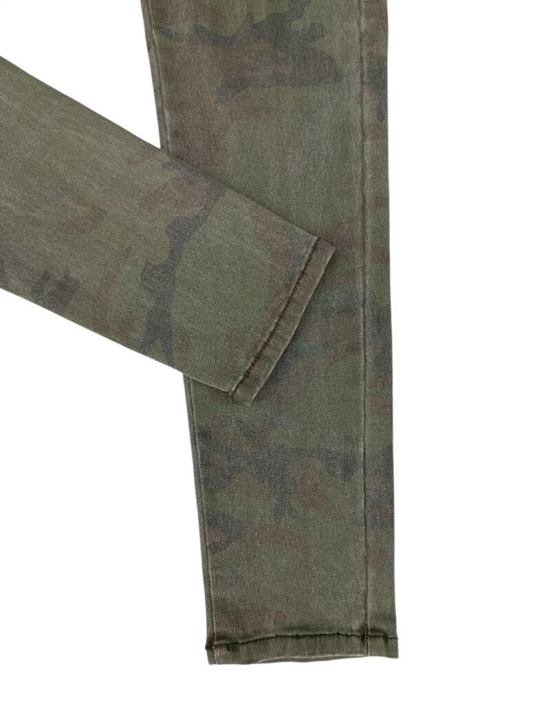 Puede incluir: Primer plano de unos vaqueros con estampado de camuflaje verde oliva. La tela presenta una mezcla de tonos verdes, marrones y negros. Los vaqueros est&aacute;n doblados, mostrando el dobladillo y la textura del material. El estilo general es informal y pr&aacute;ctico.