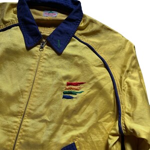 May include: A yellow satin jacket with a blue collar and blue piping. The jacket has a zipper closure and a rainbow-colored embroidered logo on the left chest. The logo says "Revolution".
