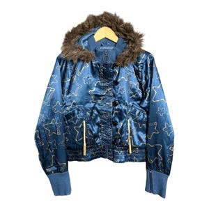 Puede incluir: Chaqueta de satén azul con estampado de estrellas y capucha forrada de piel sintética. La chaqueta tiene cierre de botones, bolsillos con cremallera y puños de canalé. Es una chaqueta estilo bomber.