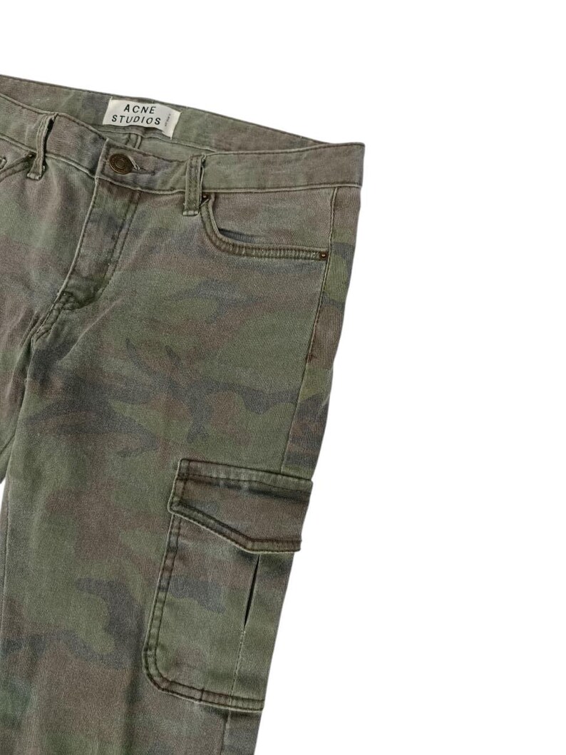 Puede incluir: Pantalones con estampado de camuflaje en tonos verdes, marrones y grises. Los pantalones tienen un bolsillo cargo y la etiqueta de la marca "ACNE STUDIOS". Tienen cierre de bot&oacute;n y cremallera, adem&aacute;s de bolsillos est&aacute;ndar.
