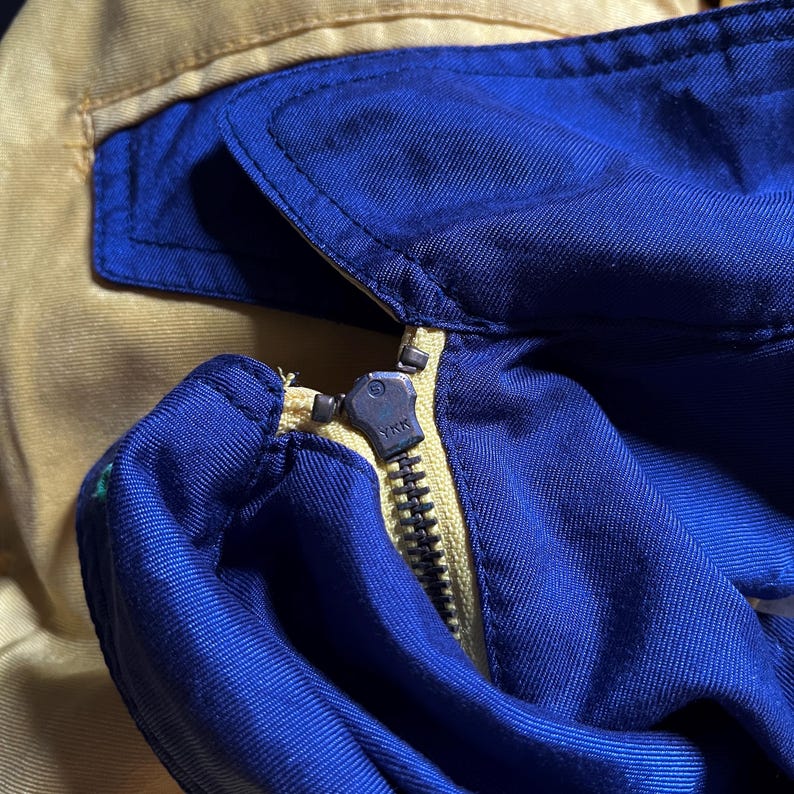 May include: Close-up of a zipper on a blue and yellow jacket. The zipper is metal and has a YKK logo. The jacket is made of a soft, textured fabric.