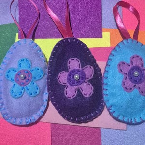 May include: Three felt Easter egg decorations in shades of purple and blue, each with a stitched flower and a pearl bead centre. They have pink ribbon loops for hanging. The background features colourful felt squares.