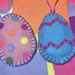 Puede incluir: Dos adornos de huevos de Pascua de fieltro. Uno es rosa con lunares coloridos y el otro es azul con un diseño en zigzag. Ambos tienen costuras moradas y una cinta para colgar. El fondo es de fieltro colorido.