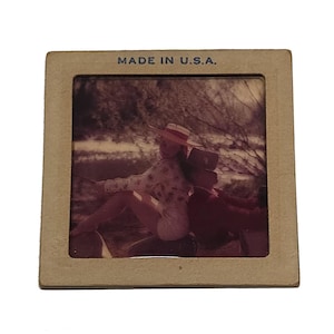 May include: A square vintage photographic slide with a tan cardboard mount featuring the text MADE IN U.S.A. at the top. The central image depicts two individuals sitting back-to-back outdoors, one wearing a wide-brimmed hat and light clothing.